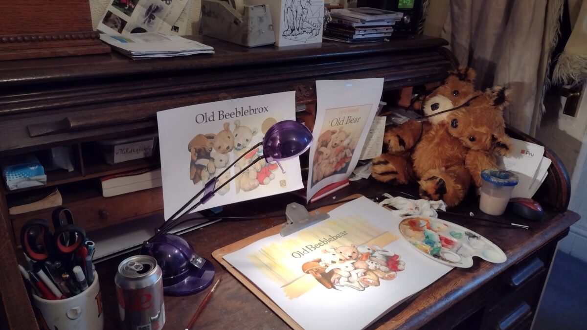 A photograph. On a wooden desk desk sits a watercolor painting, along with brushes, paint palette and reference materials. The painting has a group of stuffed toy animals. The central toy is a two-headed teddy-bear. Next to the painting is a physical brown two-headed teddy-bear.