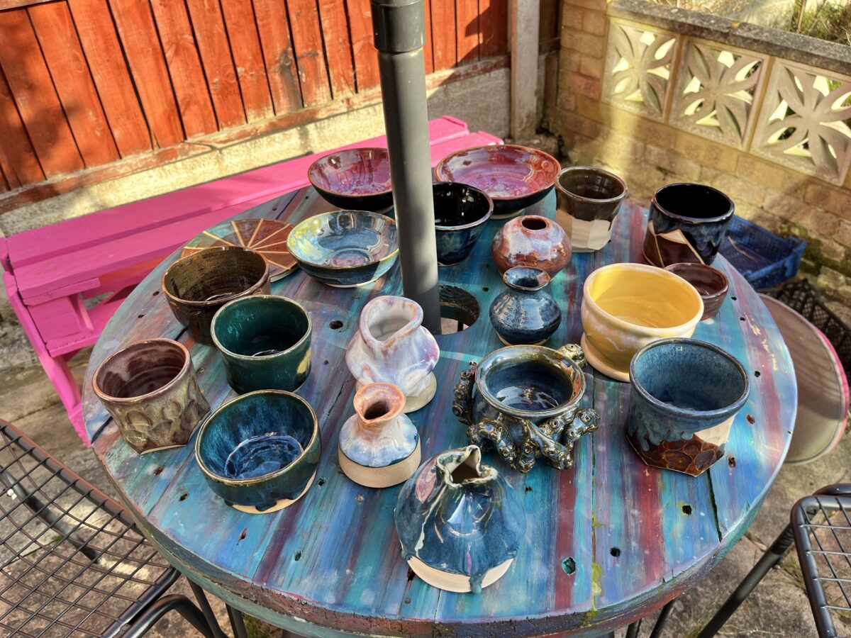 A photo of a collection of ports and reed diffusers siting on a round randbow-coloured table outside. The pots are a variety of shapes, colours and textures.