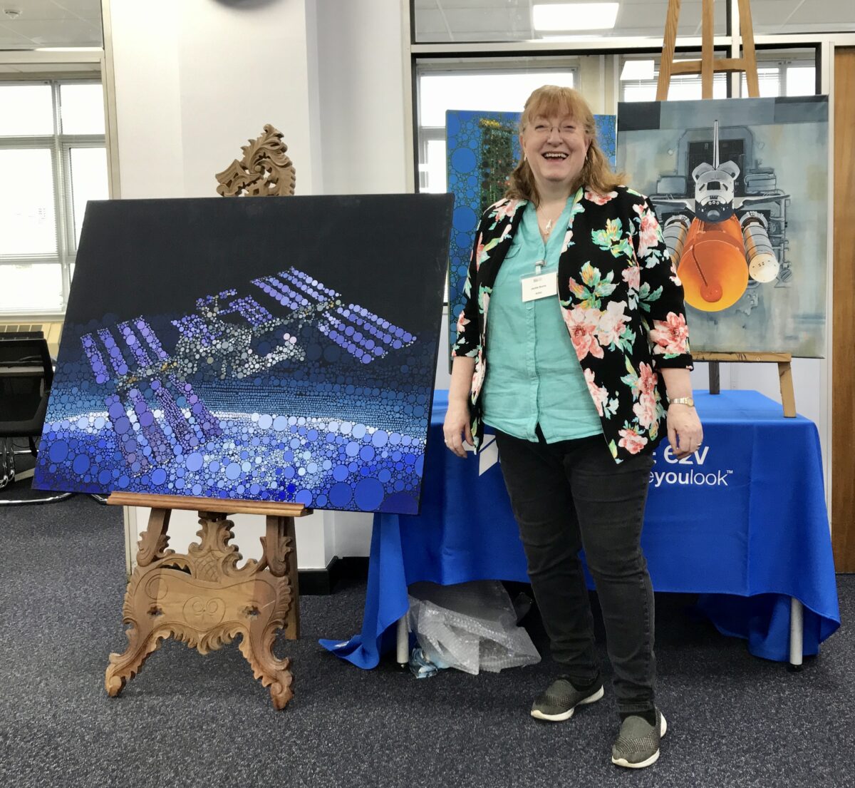 A photograph of Jackie Burns, a white woman with light hair, standing next to some of her artwork on display. To her left is an image of a space satellite made of circles in various shades of blue, purple, white and black. Behind her is an obscured image made of blue and green circles. To her right is a picture of a gray-white space shuttle coming out of an bay entrance. Below the space shuttle is a striking orange cylinder.
