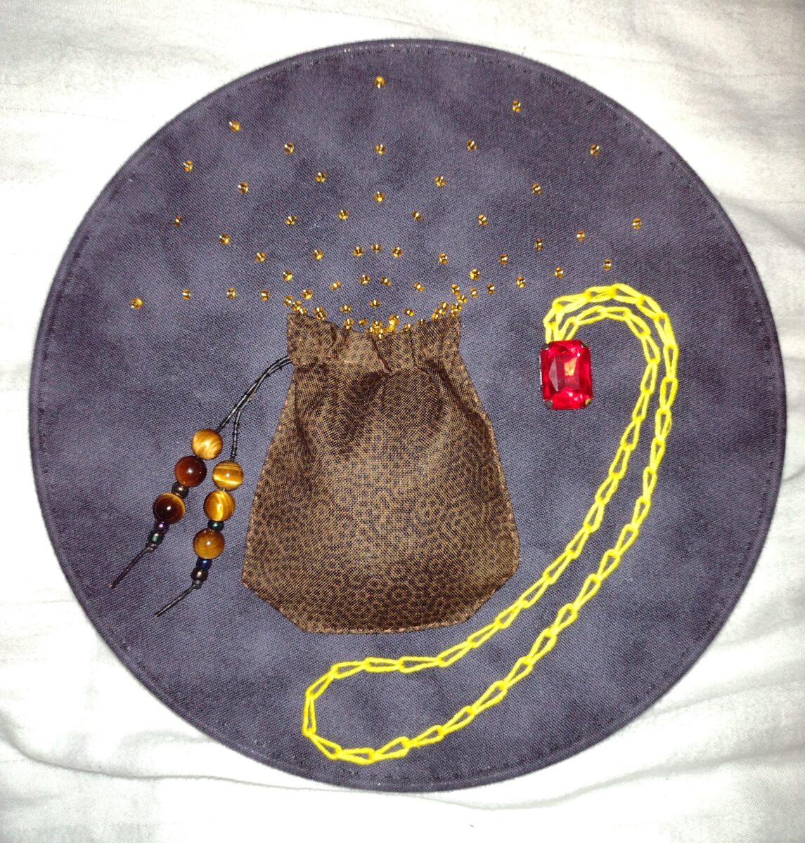 A photograph of a blue-purple fabric circle. In the centre is sown on a brown drawstring pouch with amber beads on the strings. Emanating from the pouch are arches of golden sparks. To the right is sewn on a red jewel with a golden chain.