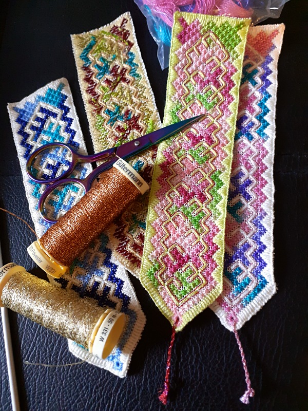 Photograph of four embroidered bookmarks with geometric patterns in different colours. On top of them sits a pair of scissors and two spools of thread.