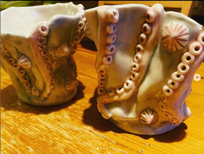 Two clay vessels that are deformed to have an organic shape. On the surface are seashells, and several tentacles stretching from top to bottom. The surface is a pale green, and the tentacles and shells a pale pink.